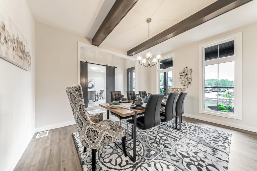 A modern dining room featuring a long wooden table set with elegant black dinnerware and surrounded by a mix of upholstered and leather chairs. The room has light-colored walls, large windows with a view of greenery, and wooden ceiling beams. A decorative rug covers the floor, and stylish light fixtures hang from the ceiling. A glimpse into another room through sliding doors is visible in the background.
