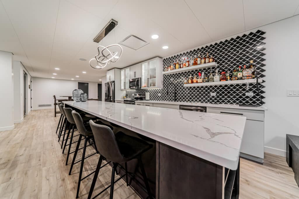 A modern kitchen showcasing a long, marble-topped island with several black stools lined up along it. The kitchen features a sleek, contemporary design with a black and white geometric backsplash. Open shelves on the wall display an assortment of liquor bottles. Above the island hangs a stylish, artistic light fixture. The flooring is light wood, providing a warm contrast to the predominantly black and white color scheme.