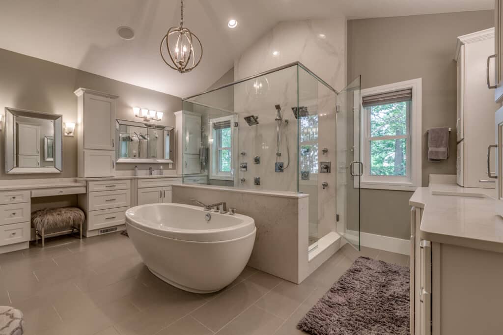 A luxury modern bathroom featuring a freestanding oval bathtub in the center, surrounded by light beige tiled flooring. To the right, there is a glass-enclosed shower with multiple showerheads. The walls are painted in a soft gray tone, and there is a large window with a view of trees. On the left, there is a double vanity with white cabinets, a large mirror, and overhead lighting. A plush gray rug and a unique modern pendant light add to the elegant decor.