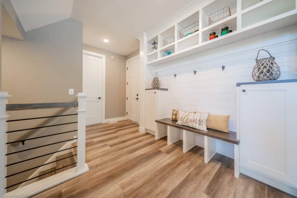 A modern entryway featuring a wooden floor with a light brown finish. On the right, there is a white shiplap wall with hooks and a built-in bench beneath it, decorated with a 'Welcome' pillow and a mustard-colored cushion. Above the bench are open shelves with decorative baskets and jars. There are cabinets on either side of the bench, and an angled stair railing is visible on the left, leading downward. The hallway has a closed white door at the end, and recessed lighting gives the space a warm glow.